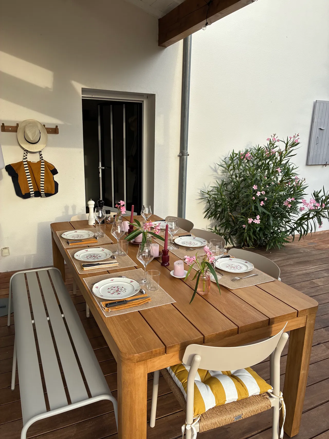 La grande table en teck dressée sous le porche : vaisselle fleurie, bouquets de laurier-rose, le banc et les chaises à coussins rayés
